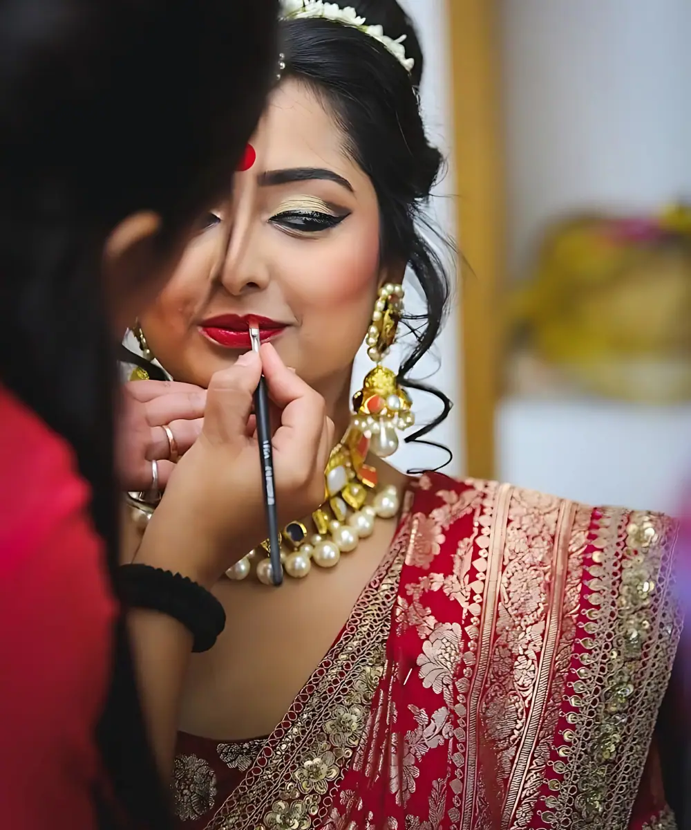 Makeup artist applying red lipstick to a bride wearing a traditional red and gold saree, pearl jewelry, and gold earrings during her bridal makeup session.